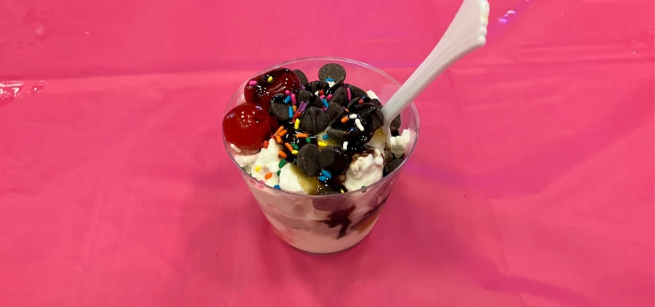 An ice cream sundae topped with cherries, sprinkles, and chocolate chips, on a pink tablecloth