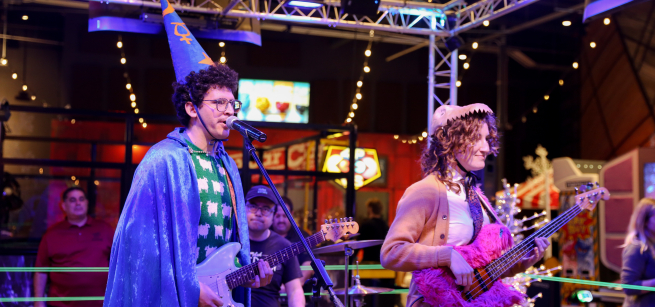 A person dressed as a wizard plays guitar while a person to their left wears a dinosaur hat, plays a fuzzy pink guitar and a drummer sits behind them in a glowing boxing ring.