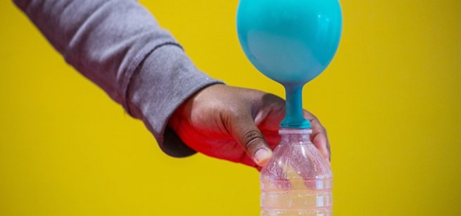 A hand holds onto a water bottle containing vinegar and baking soda with an inflated balloon at the top of the bottle. 
