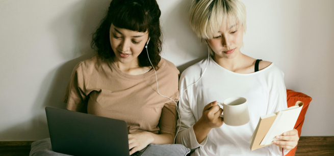Queer Asian women happily sitting next to each other. One is looking at her laptop, the other is holding a coffee cup while reading a book.