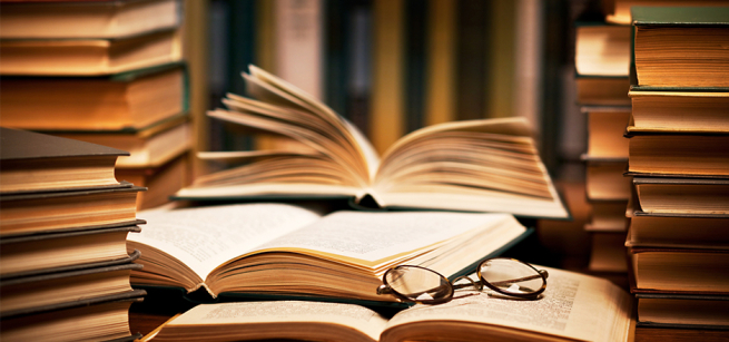 Stacks of books piled upon a desk one is open
