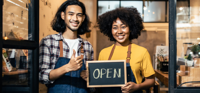 Man and woman wearing aprons stand inside a business holding Open sign