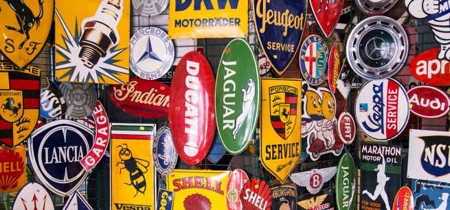 A collection of automotive signs hang in a shop, including Jaguar, Porsche, Peugeot, and Shell oil