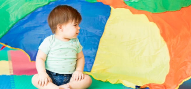 A child looks to their left while sitting on a colorful foam mat with a rainbow play parachute falling behind them. 