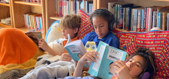 Three children, with pillows propped against bookcases, reading at a library