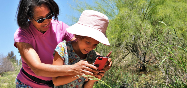 A mom helping her daughter take a photo of a plant on their cell phone.