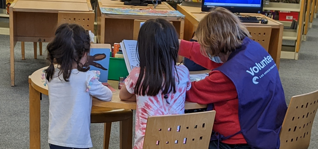 A woman wearing a volunteer vest reads to two children around a table