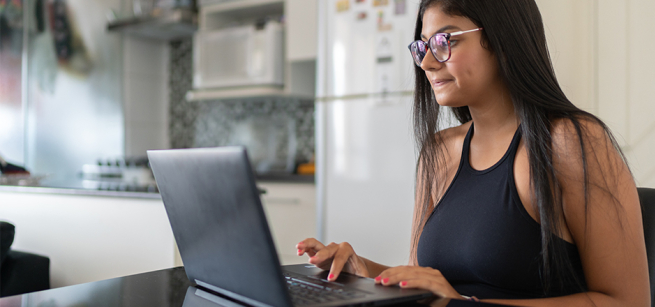 A woman with glasses and medium-length hair is seated at a desk at home, focused on using a laptop. The room has a warm and inviting atmosphere.