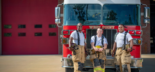 three fire fighters in uniform stand in front of a fire engine and smile at the camera