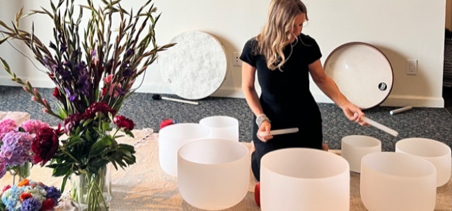 A woman in black clothing sits surrounded by crystal bowls.