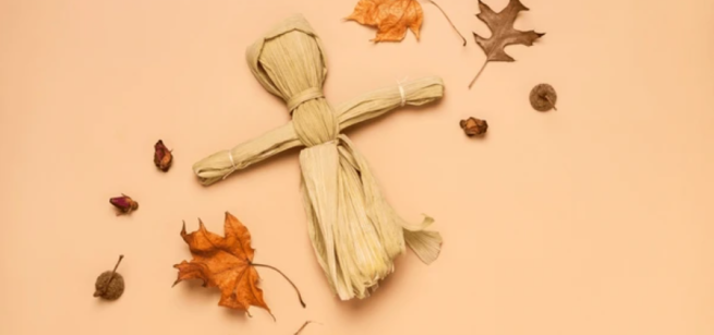corn husk doll on a peach background with fall leaves