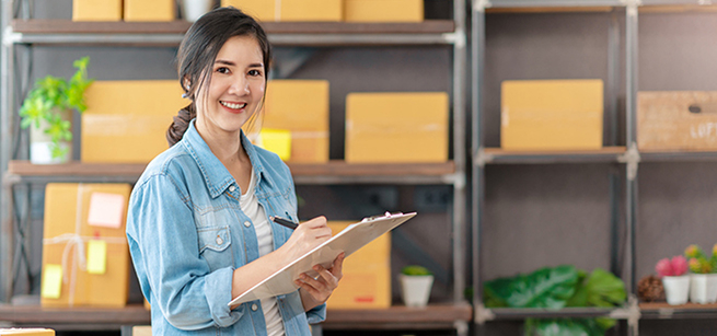 Woman business owner holding a clipboard, standing in front of a shelf with cardboard boxes.