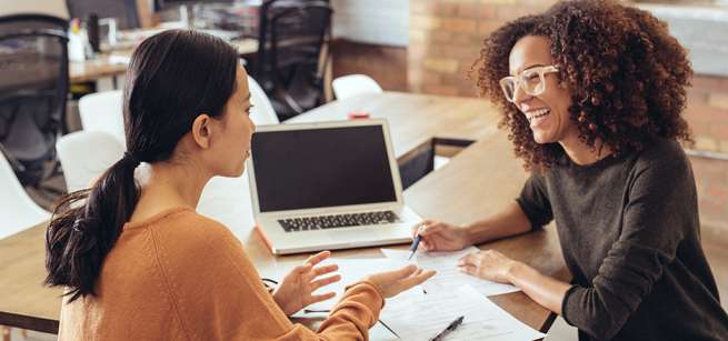 Two women discussing finances with paperwork and an open laptop in the background.