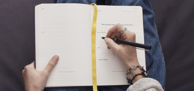 Close up of a persons hands writing in a journal on their lap.