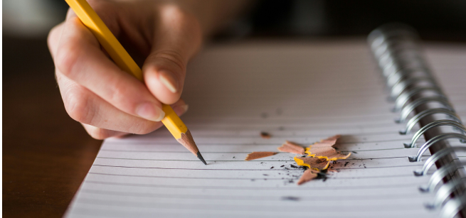 A hand holding a pencil on a blank page of a notebook with pencil shavings scattered on a small part of the page.