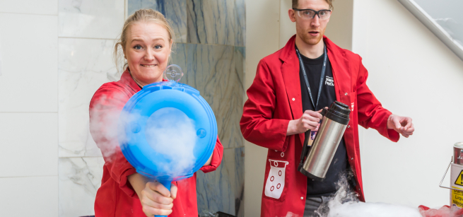 Two people in red lab coats demonstrate an experiment with dry ice