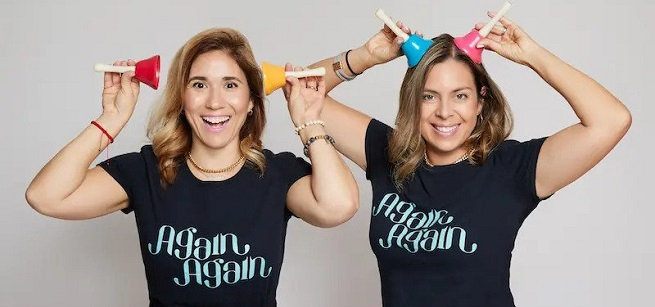 Two women holding colorful bells on their heads and wearing black T-shirts that say Again Again