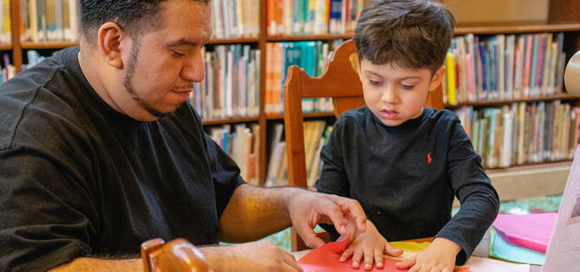 A father and son sit at a table in a library, folding colorful paper.