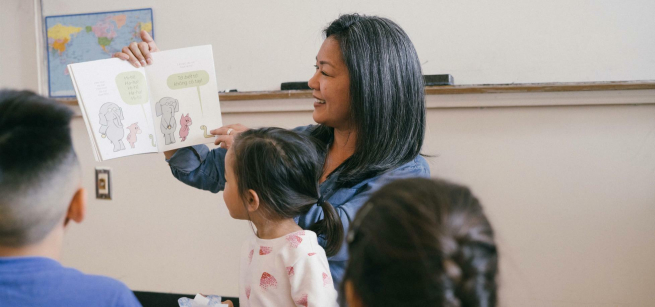 A woman holds a book open reading to children that are looking on