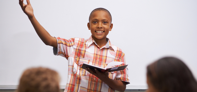 A smiling boy raises his arm as he reads a book to a classroom.