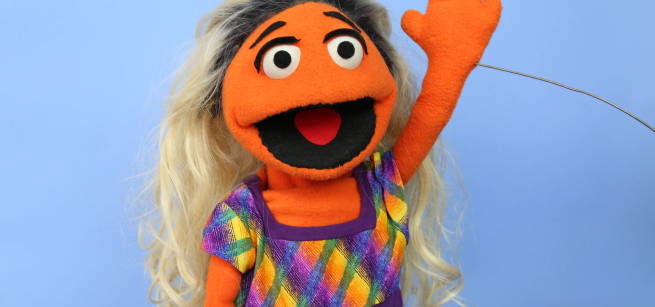 Orange Puppet girl with blond and black hair waving.