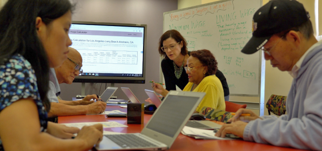 A group of adults of various ages are working collaboratively on computers in a classroom setting. 