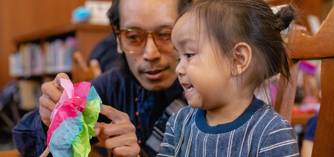 A father shows his toddler a brightly-colored toy in the library.