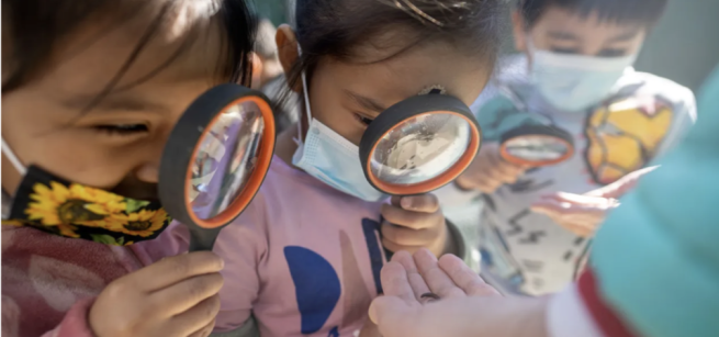3 children up close wearing face masks and looking through magnifying glasses at worms held in the palm of two hands