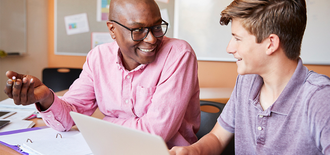 Smiling man and teenage boy talking at a desk with a laptop.