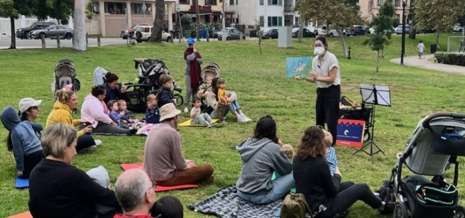 librarian reads a picturebook to a group of toddlers and adult caregivers seated on a grassy lawn in Echo Park