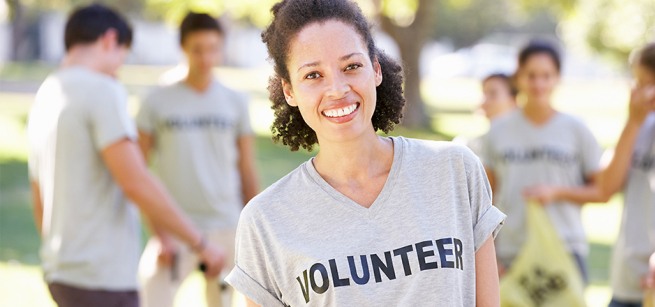 Central figure is a smiling woman wearing a grey t-shirt that says Volunteer. Behind her in a park-like environment are five people wearing the same t-shirt, interacting together.