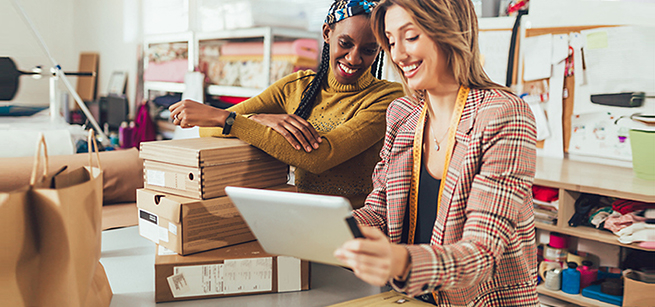 A black and a Caucasian woman are standing next two each other reviewing something in a tablet. They are in an office with boxes, materials and other tools.