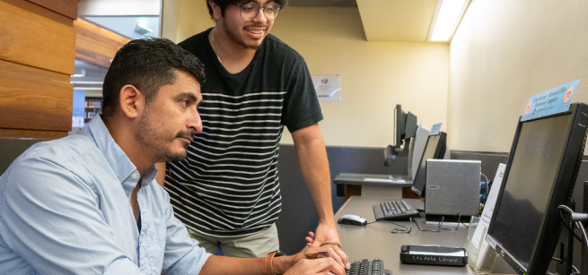 A man sits at a computer and receives assistance from another man standing next to him.
