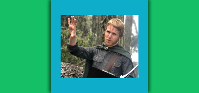 Blond man in knight costume in a wooded area.