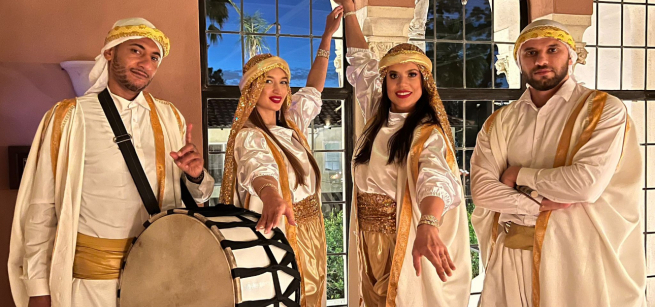 Four people in traditional attire with gold accents stand in front of arched windows; one holds a drum while others pose with hands extended.