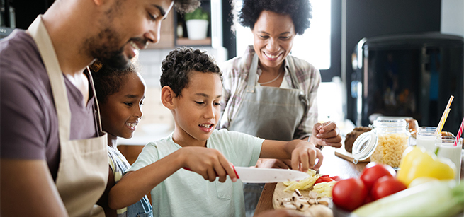 A family of four with young children are smiling while cutting vegetables and cooking together. 