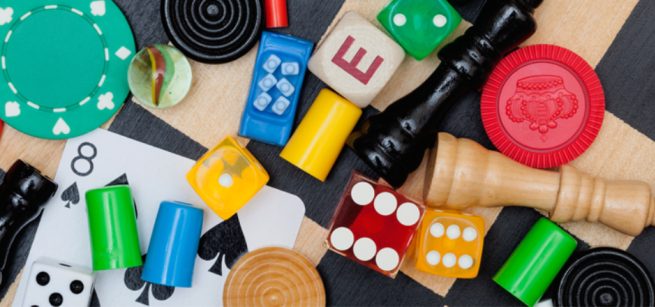A pair of dice, a letter tile, checkers and chess pieces, playing card and other game pieces scattered on a natural wood and black checkered board.