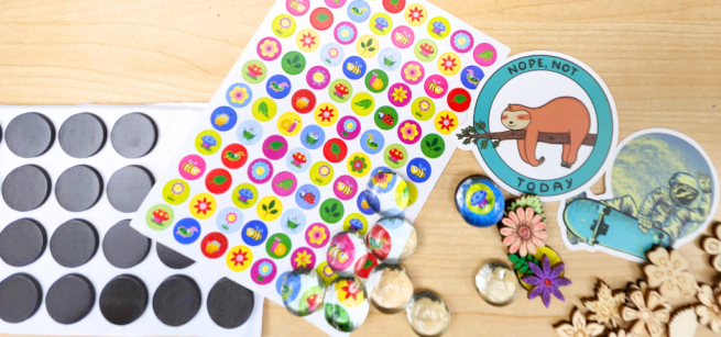 a small sheet of black magnet sticker dots, a small sheet of round colorful floral stickers, clear round glass cabochons, assorted laser cut wood flowers and leaves, and two larger stickers scattered on a light wood surface.