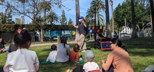 masked librarian in hat leads toddlers and caregivers seated on a grassy lawn in a fingerplay under blue sky with palm trees in the background