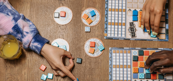 Image of 3 hands reaching from various points across a table with a board game set up. Turquoise and orange tiles are organized on different parts of the table and game board.
