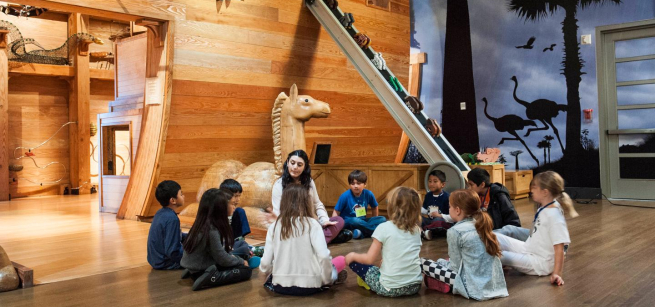 Several children and one adult interact in what looks like a stage set with a giant ark