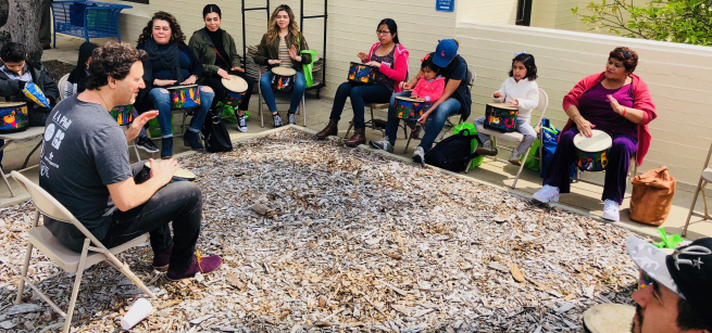 A group of twelfth drummers sitting on metal chairs with drums on their laps or between their knees, all varying in age. There’s a mix of male and female Latinos with an adult leading them. The leader is sitting on his chair in the middle-front facing the group with his drum between his knees beating a tune. This event is taking place in an outdoor patio area. Participants show eagerness and happiness and few others expressing intense concentration while trying to keep up.