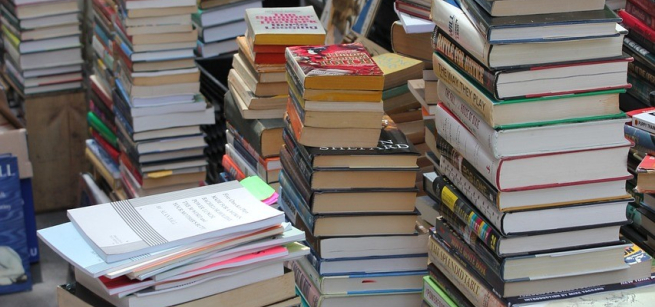 Hundred of books are stacked and the floor. They form tall towers and some are in crates.