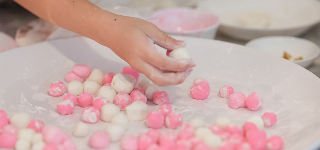 close up of a childs hand making round small dumplings. bowl filled with pink and white small round dumpligns