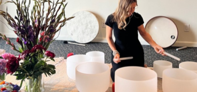A woman kneeling on the ground and surrounded by white sound bowls and a vase filled with flowers.