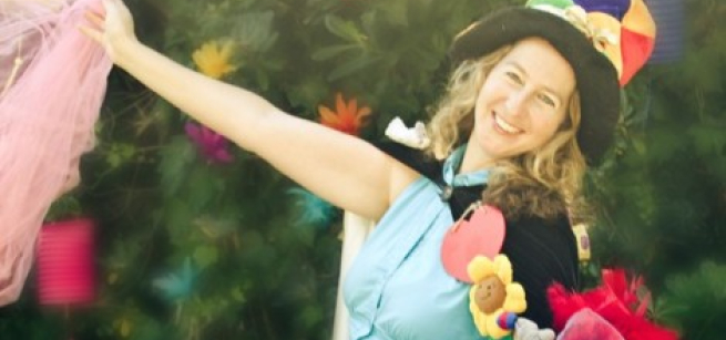 Smiling woman in a colorful hat wearing a cape and waving a sheer pink scarf
