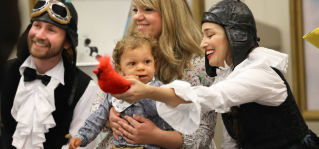 Two LA Opera Performers holding a red bird in front of a baby and his mother.
