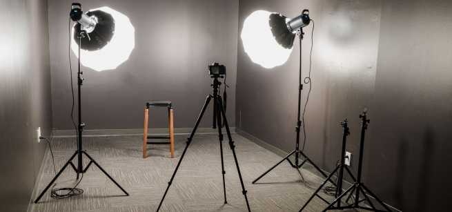A photography studio with grey walls, grey carpet, camera on tripod, large lights, and a stool