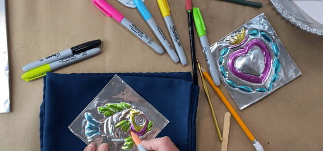 Hands working on a tin embossed art work, with a pencil and sharpies