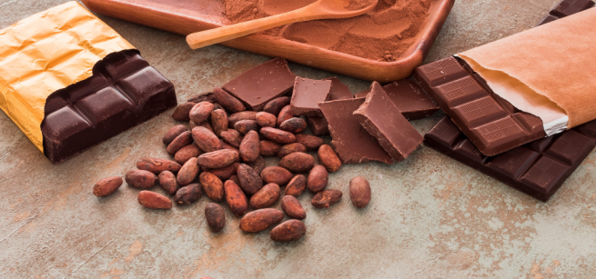 Cocoa powder in a wooden rectangular dish with a wooden spoon askew at the top, a parially opened dark chocolate bar in gold foil on the left, scattered roasted cacoa beans, at the center, and an opened milk chocolate bar wrapped in paper with a dark chocolate bar underneath. All items are over a gray surface.